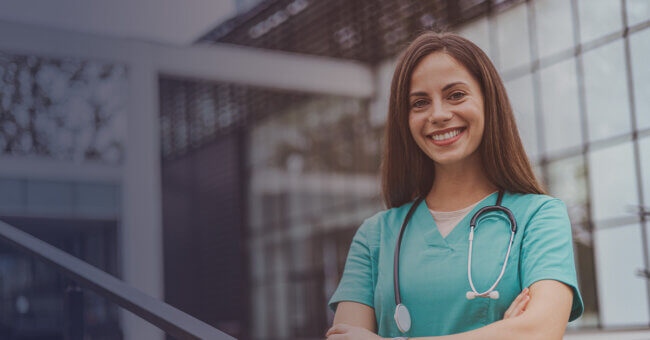Smiling female healthcare professional in scrubs with a stethoscope, standing outdoors near a modern building.